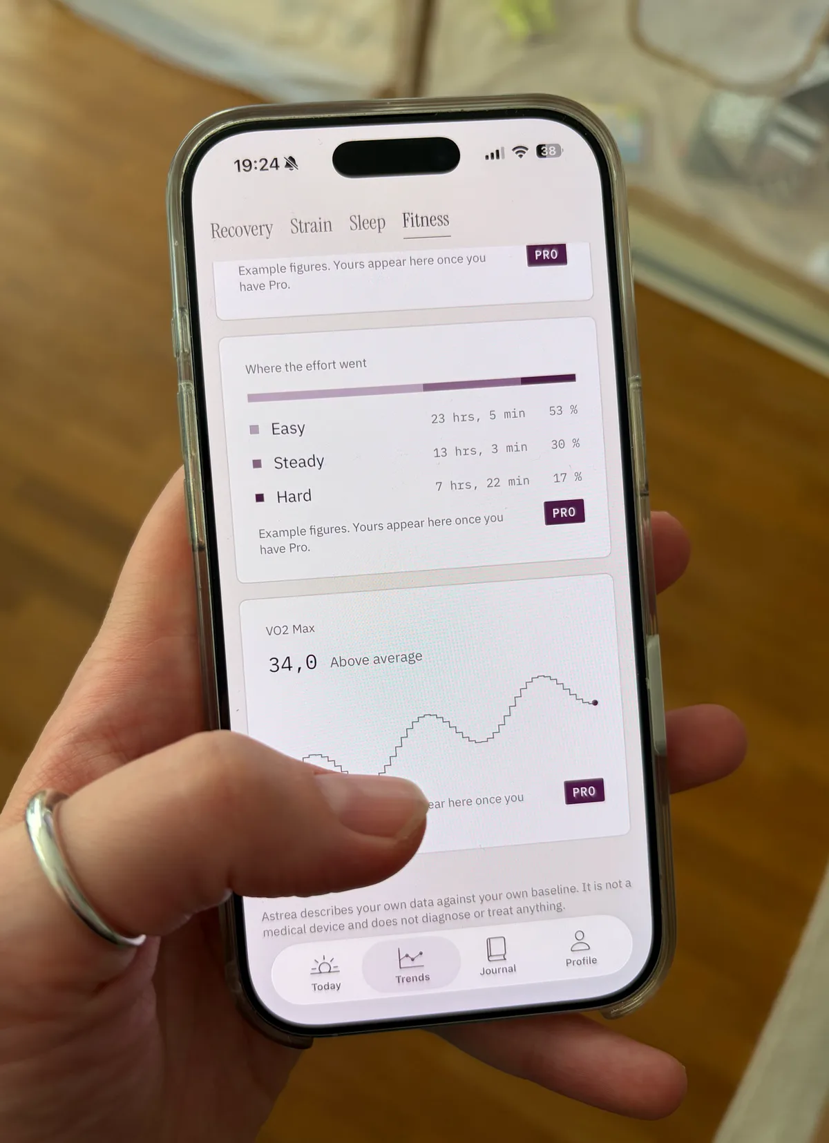 A hand holding an iPhone showing Astrea's Trends tab on the Fitness segment: a bar splitting training hours into easy, steady and hard, and a VO2 Max reading of 34.0 over a ninety-day line.